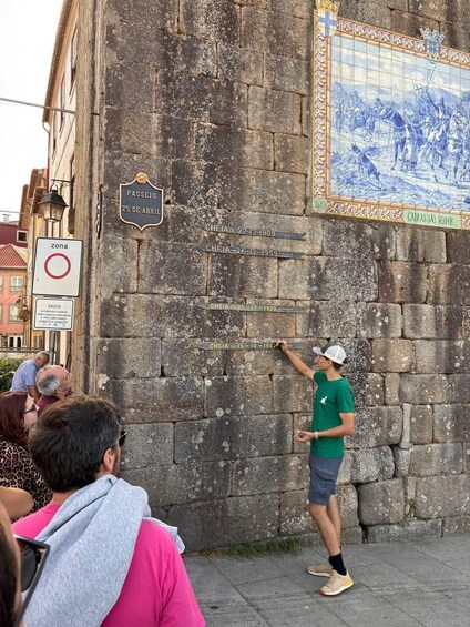 A man pointing at a wall with historical plaques and a tiled mural. 2