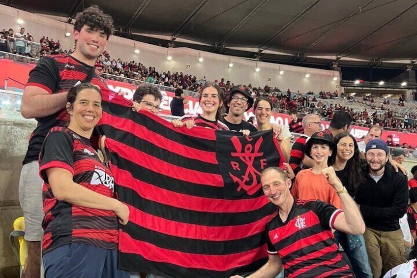 Un grupo de personas en un estadio vistiendo camisetas a rayas negras y rojas, sosteniendo una gran bandera negra y roja con un logotipo. 1