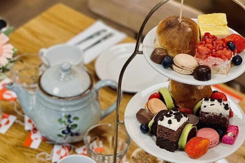 A tea set with a teapot and cups, a tiered dessert stand with pastries and fruit, and a floral tablecloth. 2