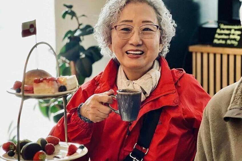 A person in a red jacket holding a mug, with a tiered tray of food and a small flag in the background. 1