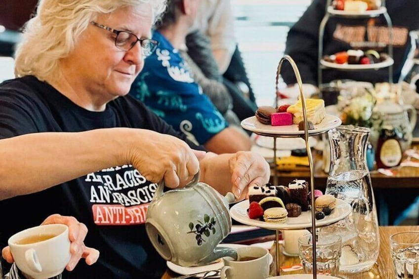 A person pouring tea from a teapot into a cup at a table with a tiered dessert stand and a pitcher. 5