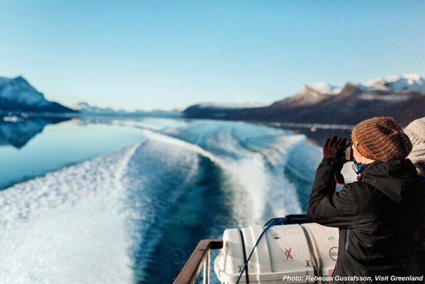A person taking a photo of a boat on a calm water body with mountains in the background. 1