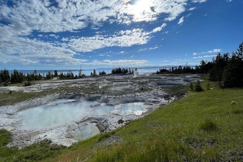 Ett geotermiskt område med bubblande lerpölar, omgivet av skogbeklädda kullar och en klar, blå himmel. 2