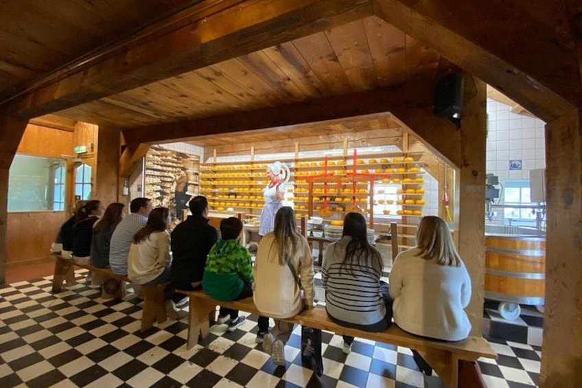 Een groep mensen zit op houten banken in een gebouw met een geruite vloer, en kijkt naar iemand in een witte jurk achter een toonbank met gele spullen. 3