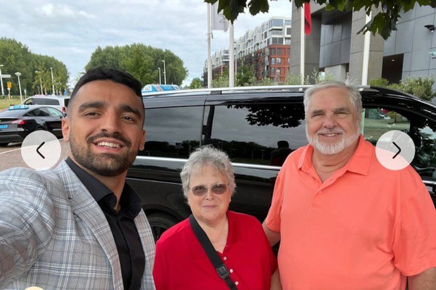 Drie personen staan voor een zwarte auto, met een gebouw op de achtergrond. 2