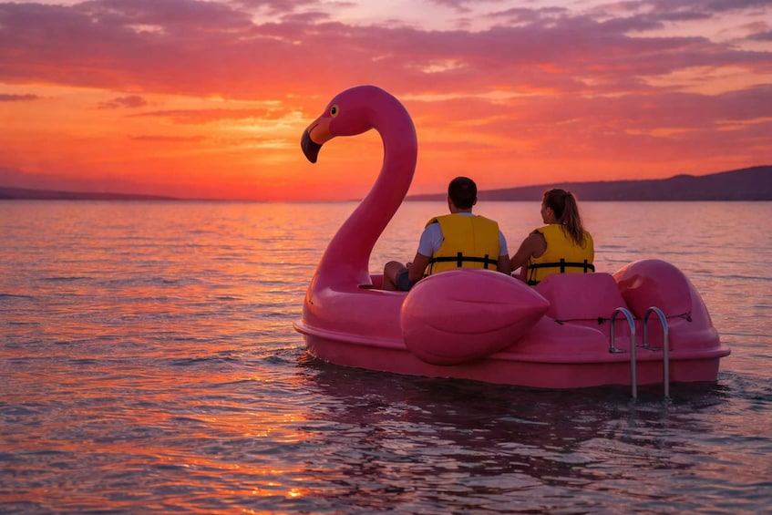 Two people on a pink flamingo-shaped raft in calm water during sunset. 1