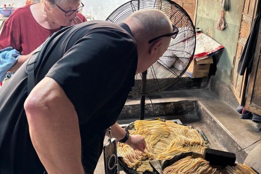 A person working with long, thin objects on a table, possibly food preparation, in a narrow space with a fan and boxes in the background. 2