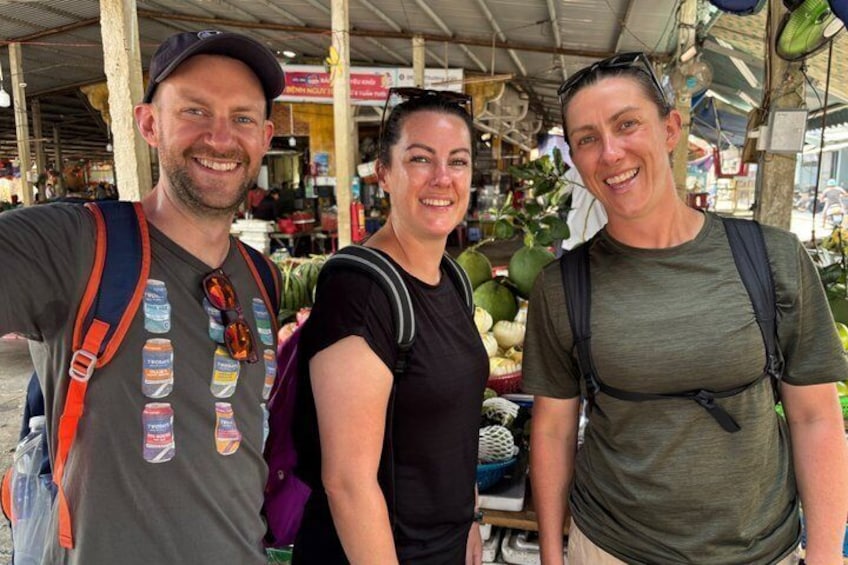 Three people standing outdoors with backpacks, a market stall with fruits and vegetables in the background. 5