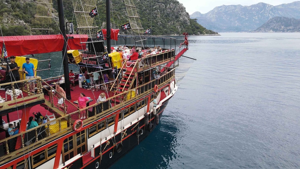 A red and white boat with a pirate-themed deck, passengers, and a mountainous backdrop. 3