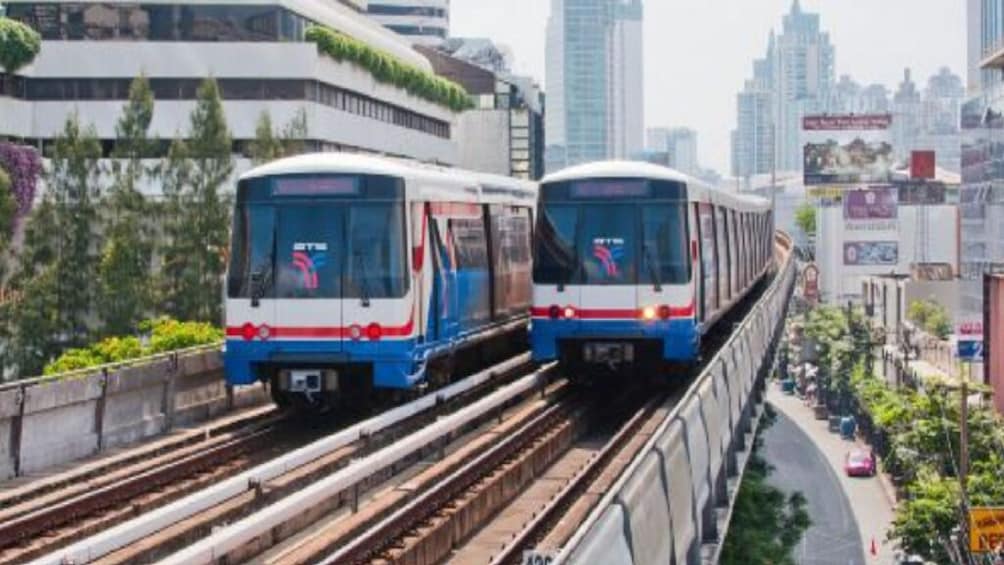 Two trains on elevated tracks in an urban setting with buildings and greenery in the background. 1