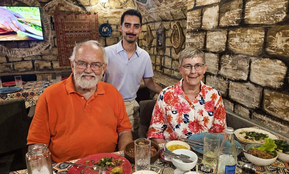Three people seated at a table with various dishes, in a room with stone walls and a television screen. 2