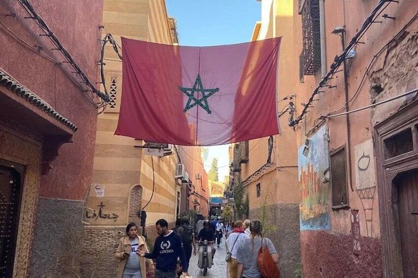 A narrow street with buildings on either side, a red flag with a green star, and people walking. 2