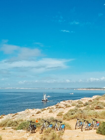 A coastal landscape with a row of bicycles parked on a sandy area, a sailboat in the distance, and a clear blue sky. 4
