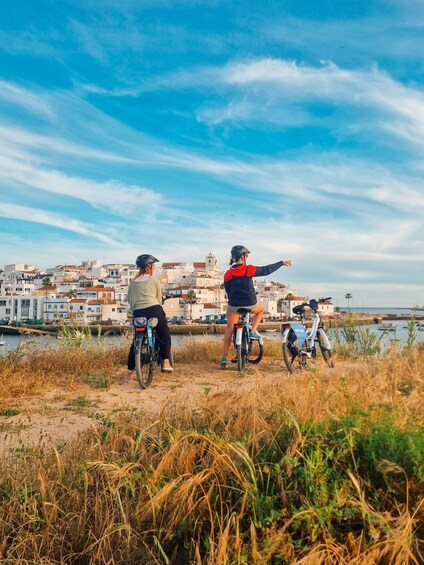Two people on bicycles, one pointing, with a coastal town and water in the background. 1