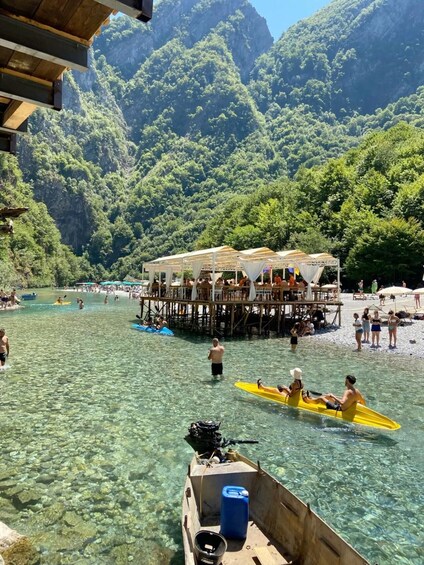 A clear water body with people kayaking, a wooden dock with a covered seating area, and lush green mountains in the background. 2