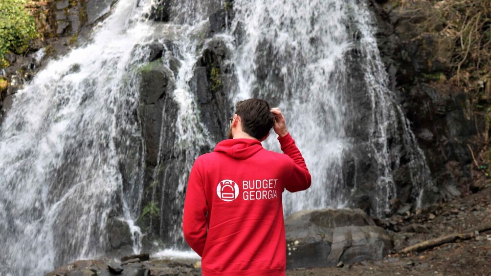 A person in a red hoodie with 'BUDGET GEORGIA' text standing in front of a waterfall. 1