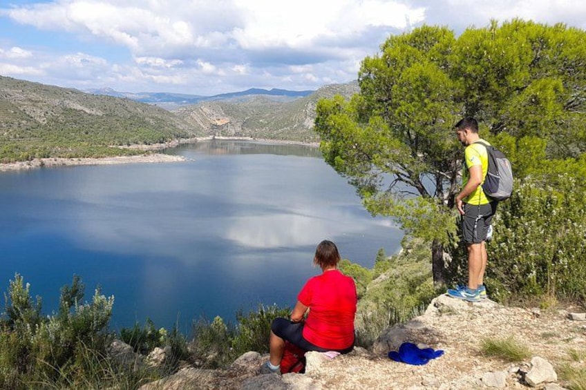 A lake surrounded by hills, a person sitting on a rock, and another standing with a backpack. 5