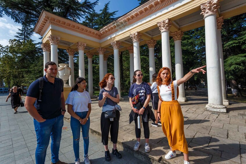 A group of people standing under a classical-style pavilion with columns, surrounded by trees. 3