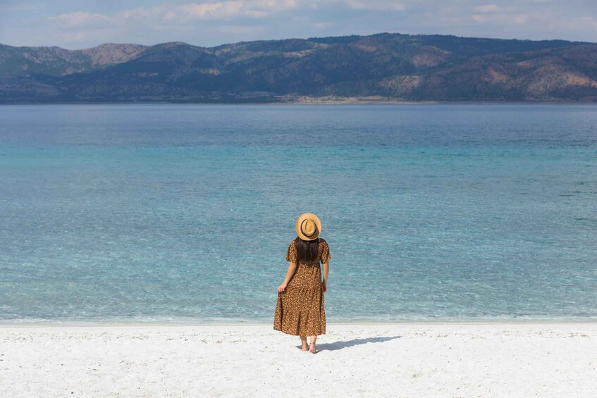A person standing on a beach facing a calm sea with clear water and distant mountains. 4