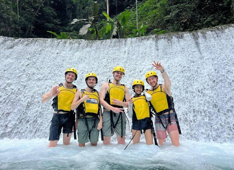 A group of people in yellow helmets and life vests standing in front of a waterfall. 1