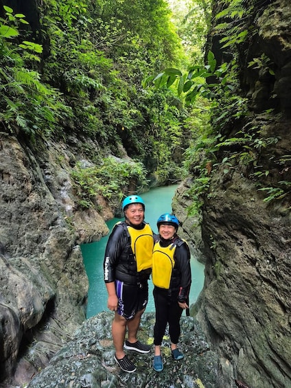 Two people standing on a rocky ledge with a narrow water channel below, surrounded by dense greenery. 4