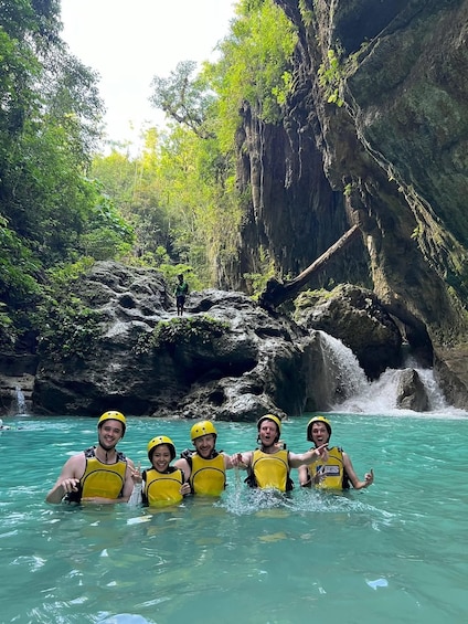 A group of people in yellow life vests and helmets in a turquoise river surrounded by rocky cliffs and greenery. 5