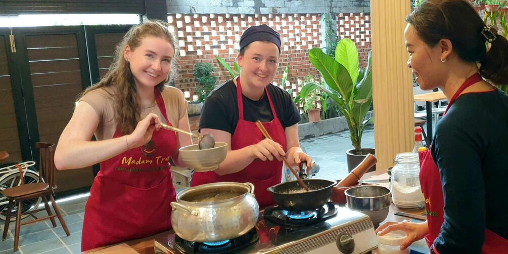 Three people cooking in a kitchen, wearing aprons, with a pot on a stove and various cooking utensils. 1