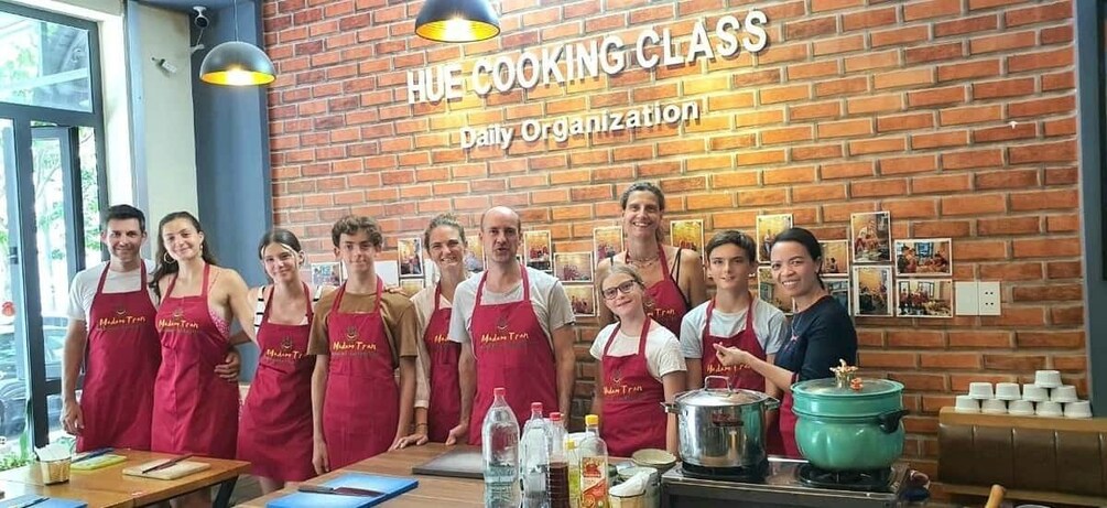 A group of people in aprons standing in a kitchen with a brick wall and a sign that reads 'HUE COOKING CLASS Daily Organization.' 2