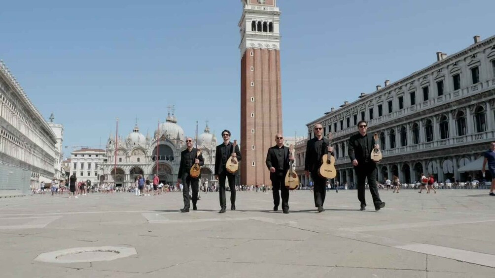 A group of musicians in a square with historic buildings and a prominent clock tower in the background. 3