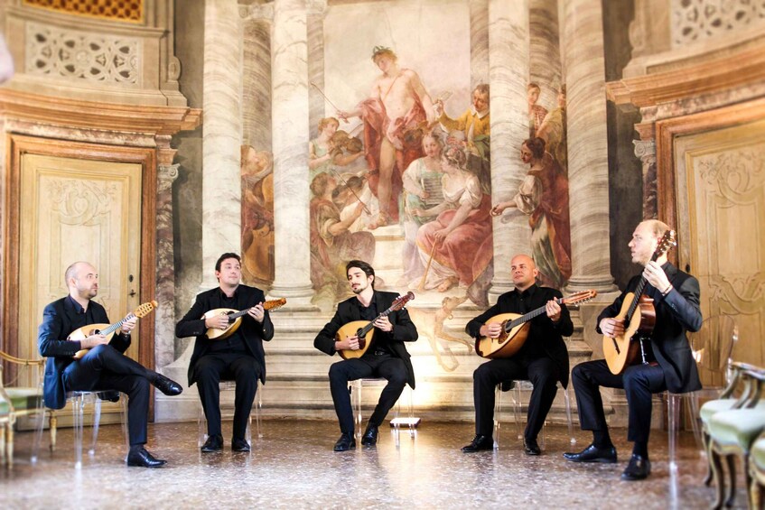 A group of musicians seated on chairs, playing string instruments in a room with ornate columns and a classical painting on the wall. 4