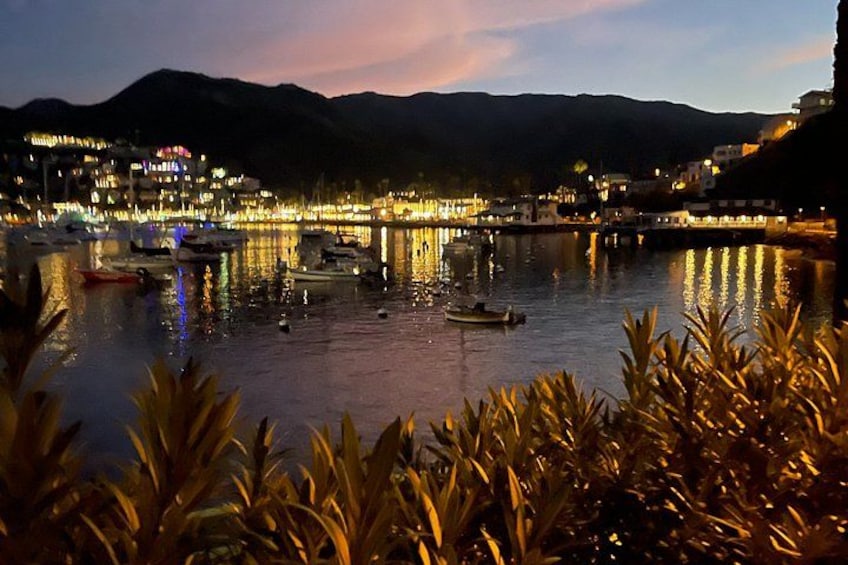 Une scène côtière nocturne avec des bâtiments illuminés, des bateaux sur l’eau et des montagnes en arrière-plan. 3