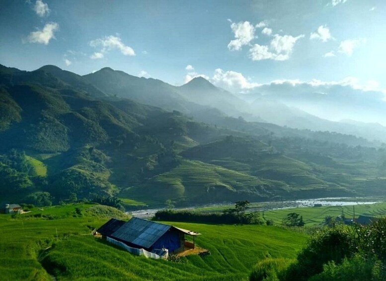 A lush green valley with terraced fields, a small house with a blue roof, and mountains in the background. 1