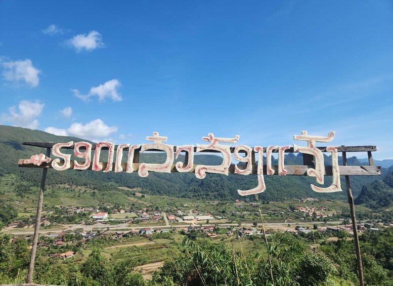 A sign with text in a non-Latin script, set against a backdrop of a green valley with scattered houses and hills. 4