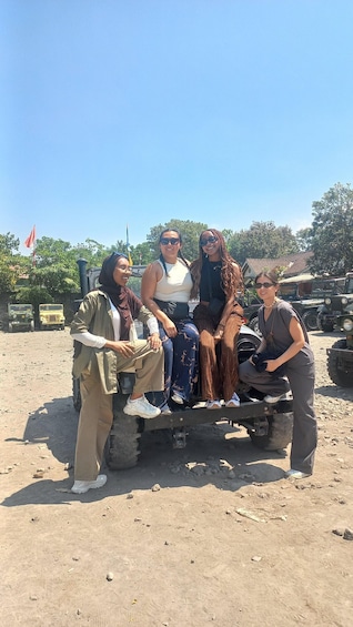 Four people sitting on a vehicle in a dusty outdoor area with military vehicles in the background. 4