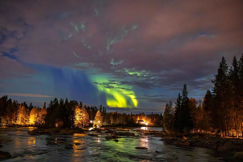 A river at night with aurora borealis, trees, and distant lights. 1