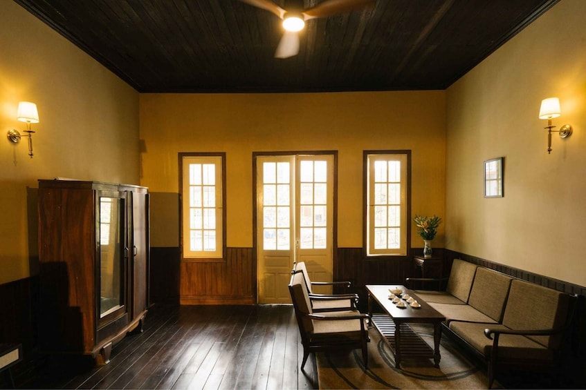 A room with wooden floors, a ceiling fan, a table with chairs, and three windows with wooden frames. 1