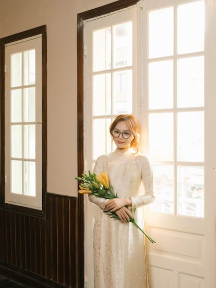 A person in a white lace dress holding a bouquet of flowers, standing in front of a door with a window. 5