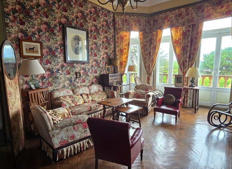 A vintage living room with floral wallpaper, antique furniture, and a view of greenery outside. 5