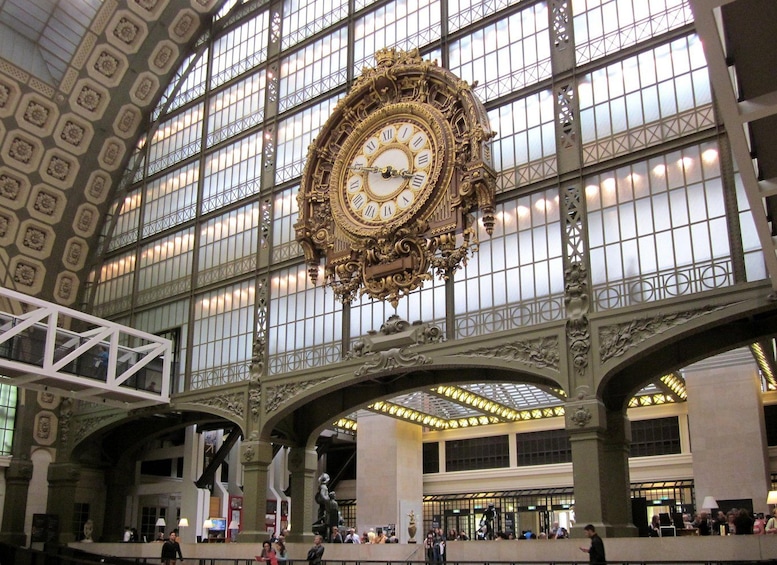 A grand, ornate clock in a spacious train station with high ceilings and large windows. 1