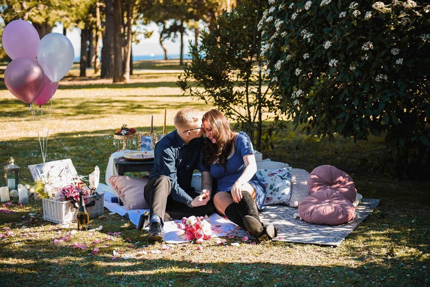 A couple sitting on a blanket with a picnic setup, surrounded by greenery and balloons. 3