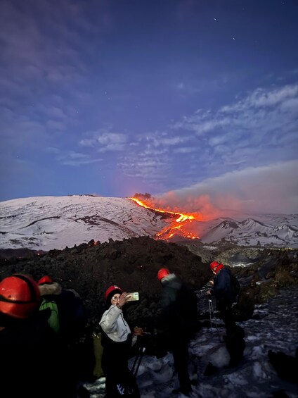 A group of people observing a volcanic eruption with lava flowing down a snow-covered mountain. 2