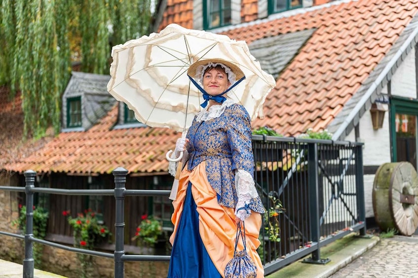 A person in historical costume holding an umbrella, standing in front of a building with a tiled roof. 1