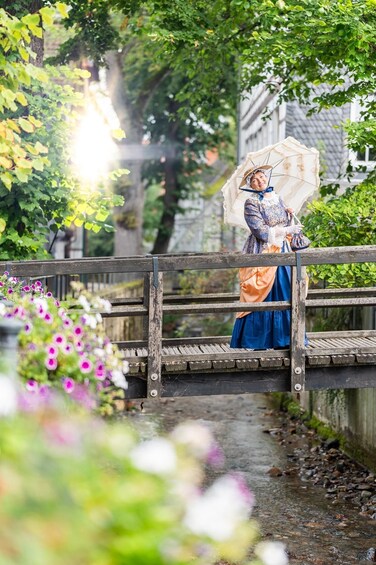 A person in historical attire standing on a wooden bridge with a floral garden in the foreground. 2