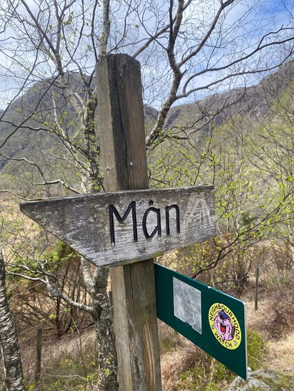 A wooden signpost with 'Man' carved on it, surrounded by trees and mountains. 5