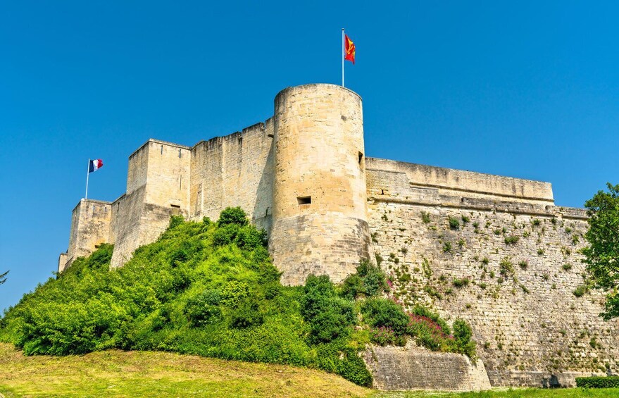 A historic stone castle with flags, lush greenery, and a clear blue sky. 2