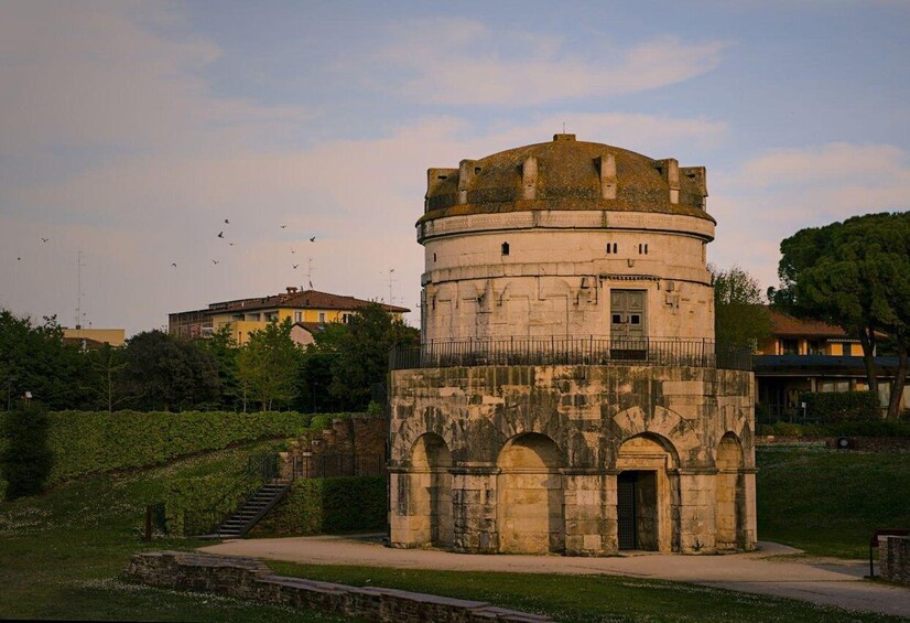 A historic tower with a circular base and arched windows, surrounded by greenery and modern buildings. 3