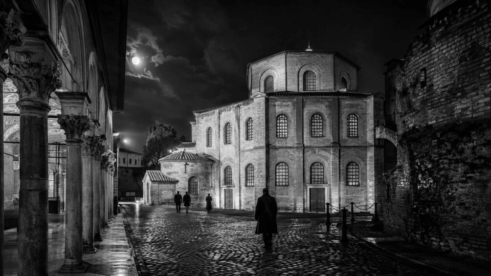 A historic stone building with arched windows and a dome, a cobblestone street, and a person walking. 1