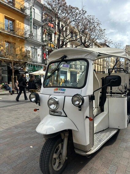 A white three-wheeled vehicle parked on a city street with pedestrians in the background. 3