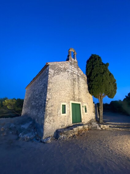 A rustic stone building with a green door and windows, surrounded by a dirt ground and a single tree. 5