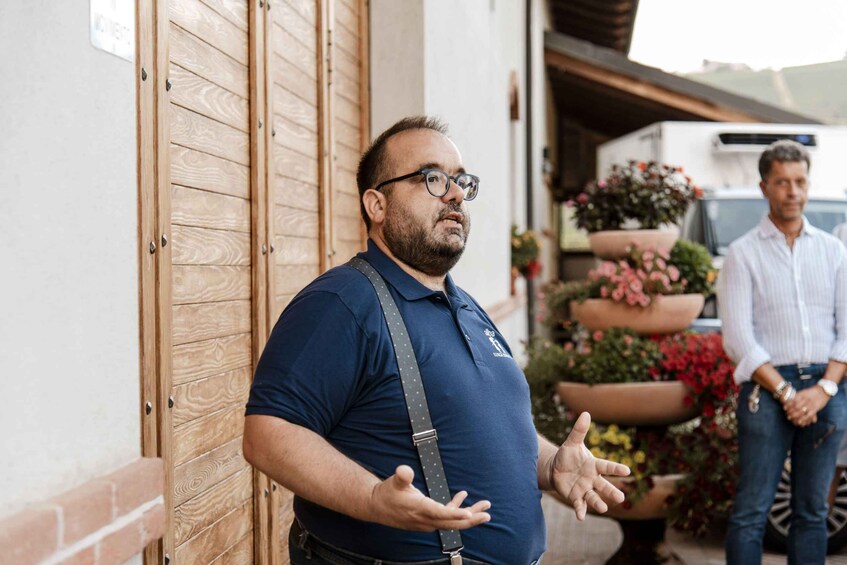 A man in a blue shirt gesturing, standing by a wooden door with a brick wall, and a truck with flowers in the background. 1